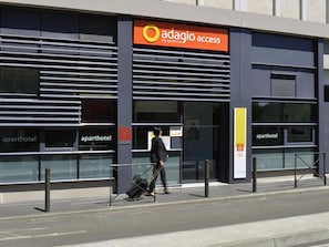 Un edificio moderno con la señalización 'adagio access city aparthotel', una persona con equipaje y una entrada despejada.