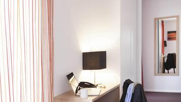 A hotel room with a desk, a lamp, a laptop, a phone, a mug, and a coat hanging on the wall.