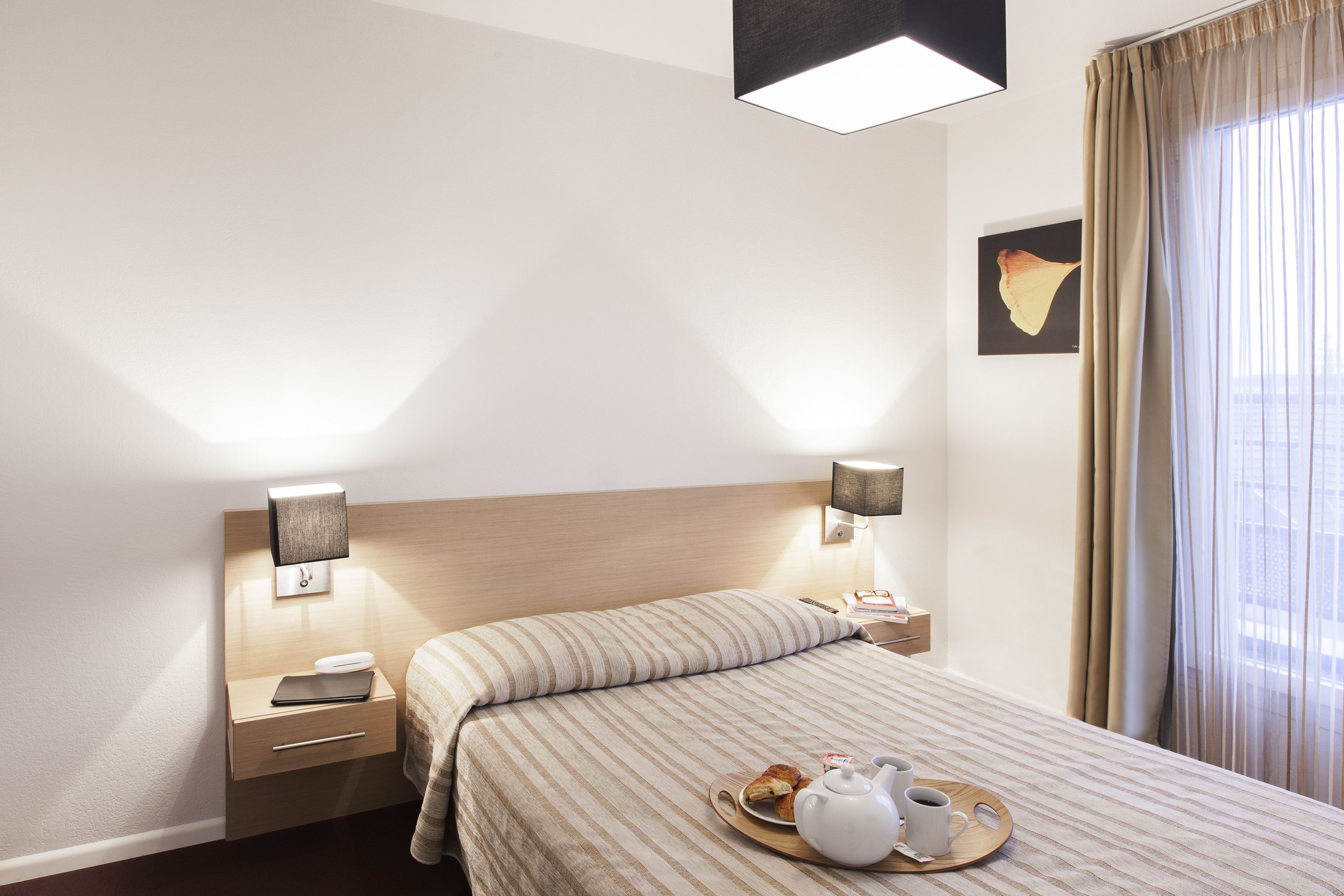 A hotel room with a bed, bedside tables, a nightstand, a tray with breakfast items, and a window with curtains.