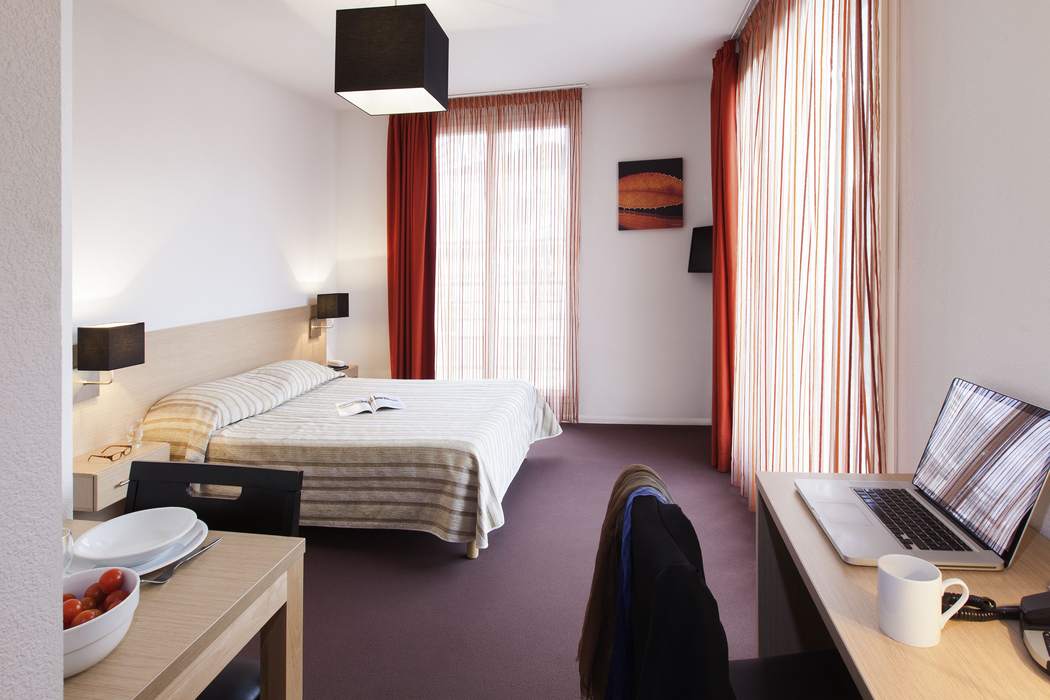 A hotel room with a bed, a desk with a laptop, a bowl of fruit, and a window with curtains.
