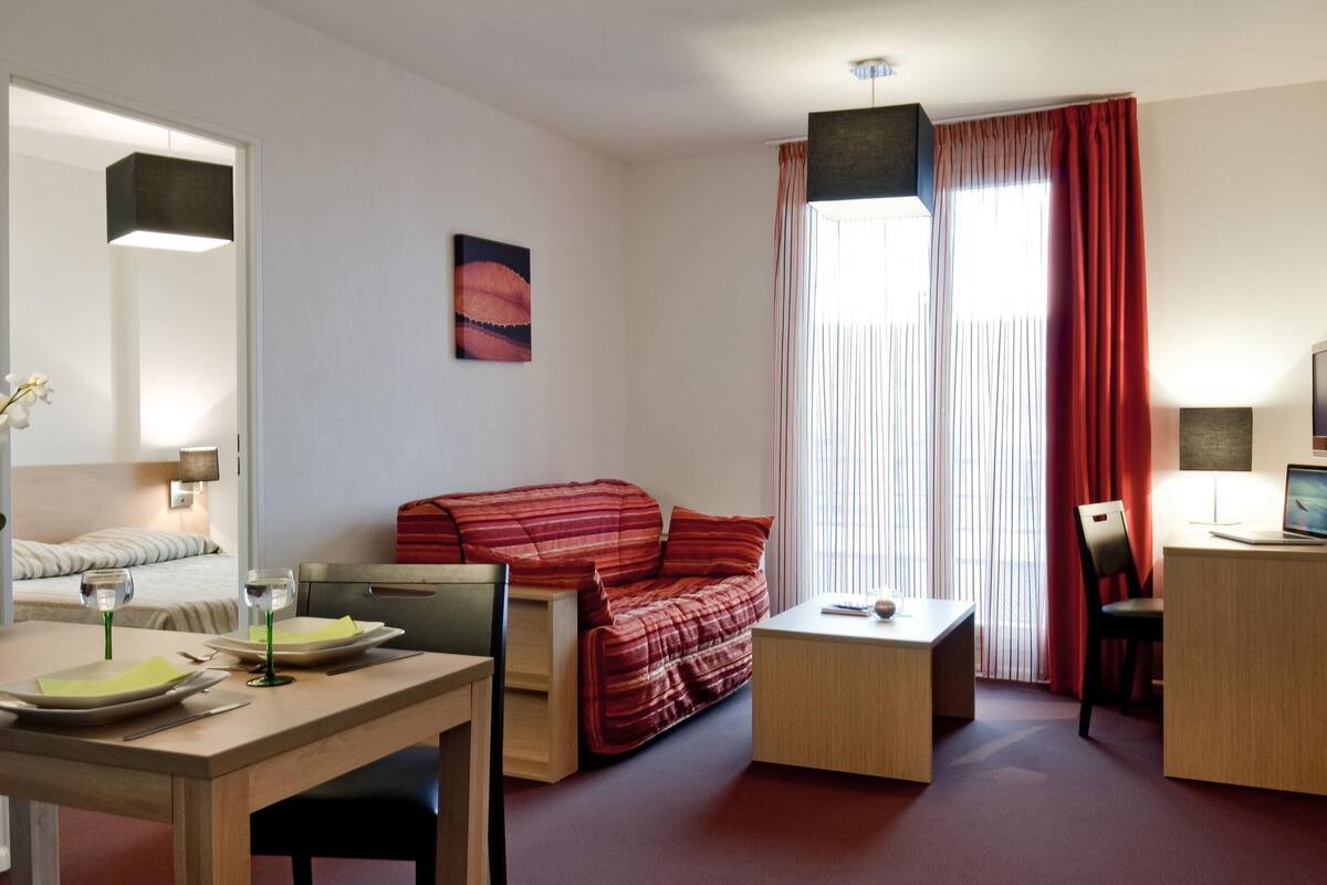 Una habitación con una cama, un sofá rojo, una mesa de madera con una computadora portátil, una mesita de centro pequeña y un escritorio con una computadora.