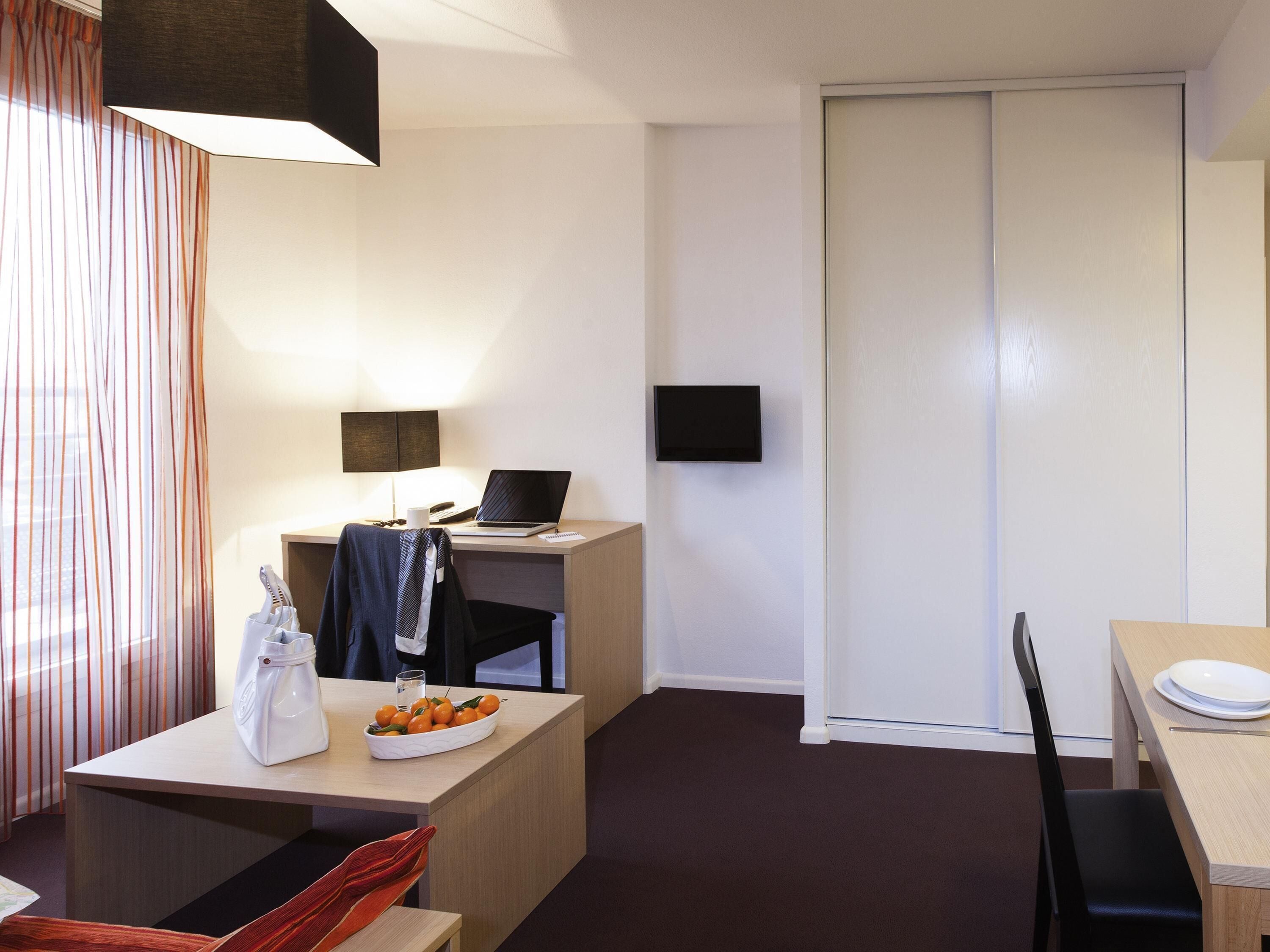 A hotel room with a desk, a laptop, a chair, a dining table with a plate and a bowl of fruit, a coat rack, and a sliding door.