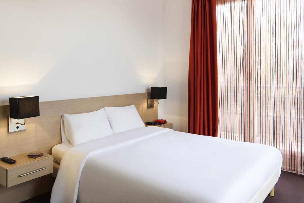 Una habitación de hotel con una cama, mesitas de noche, lámparas y un ventanal con cortinas.
