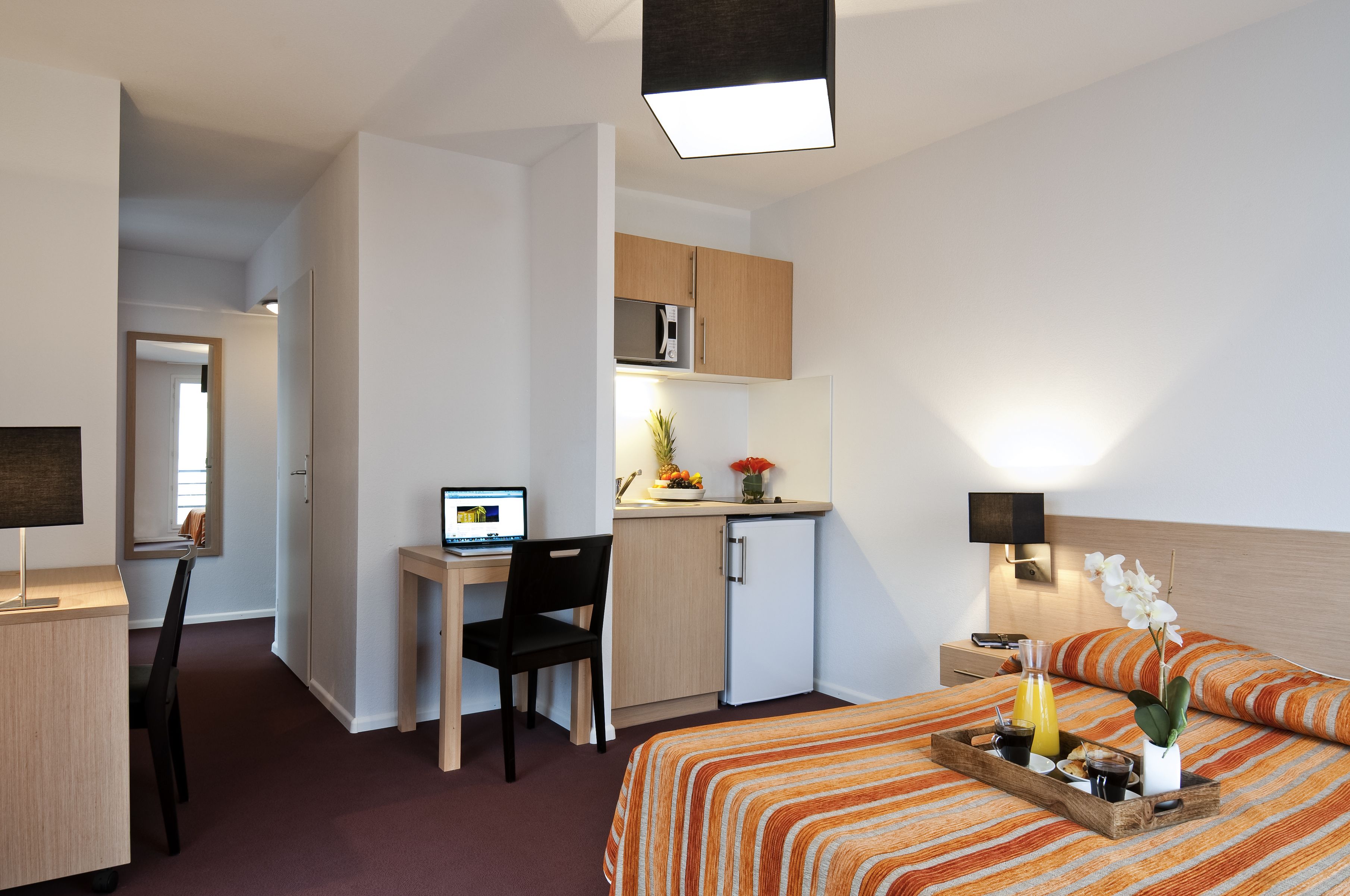 Une chambre d’hôtel compacte comprenant un lit, un bureau avec un ordinateur, une petite kitchenette et une table de chevet avec une lampe.