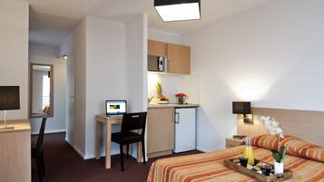 Une chambre d’hôtel compacte comprenant un lit, un bureau avec un ordinateur, une petite kitchenette et une table de chevet avec une lampe.