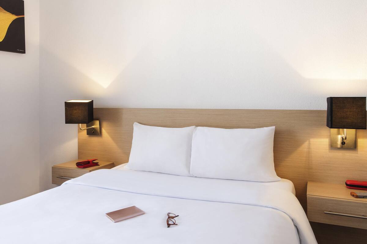 Una cama bien hecha con ropa blanca, una mesita de noche con una lámpara, y un objeto rojo pequeño sobre la mesa.