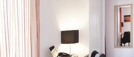 A hotel room with a desk, a lamp, a mirror, and a coat rack.