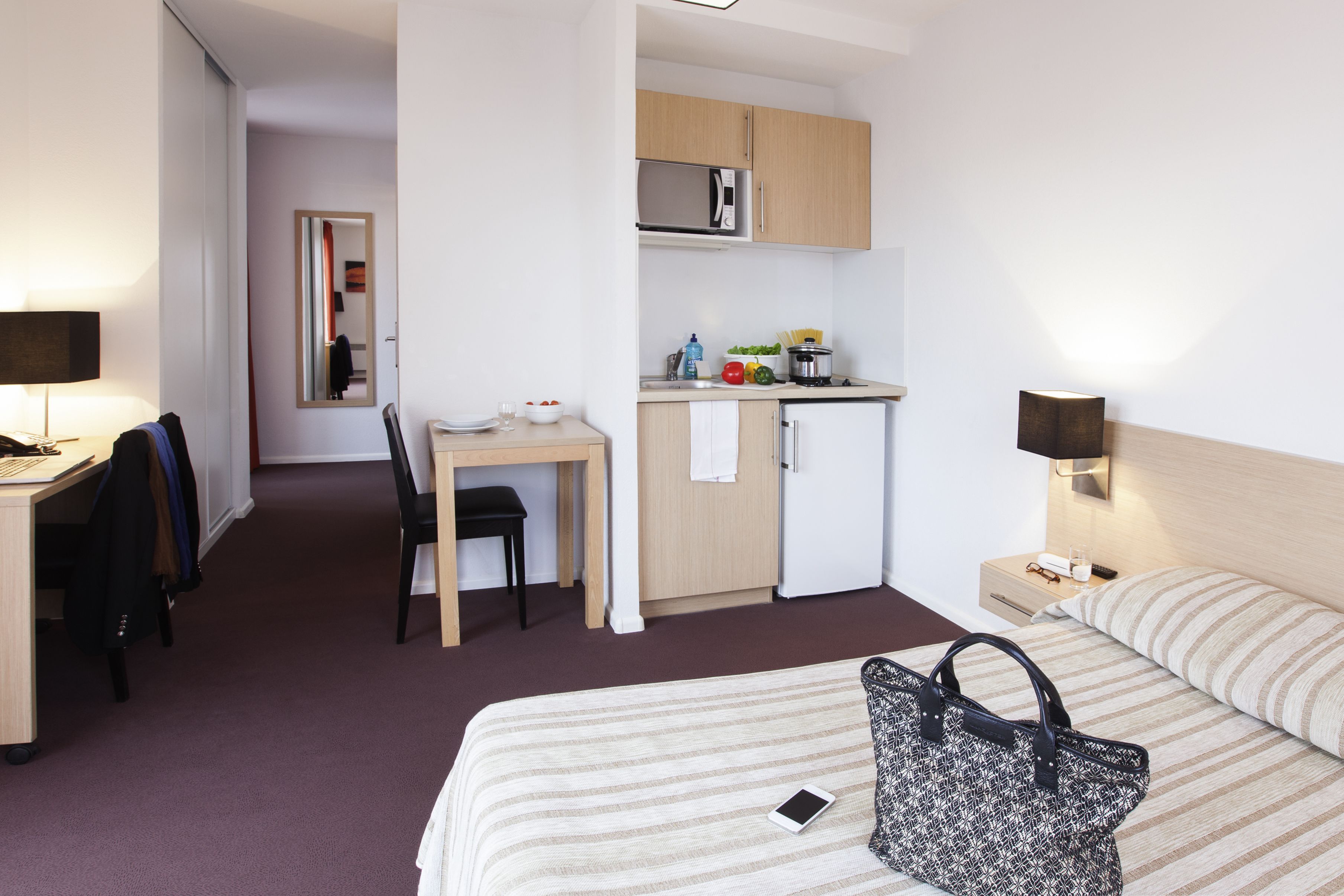 A hotel room with a bed, a desk, a chair, a microwave, a small kitchen area, and a nightstand with a lamp.