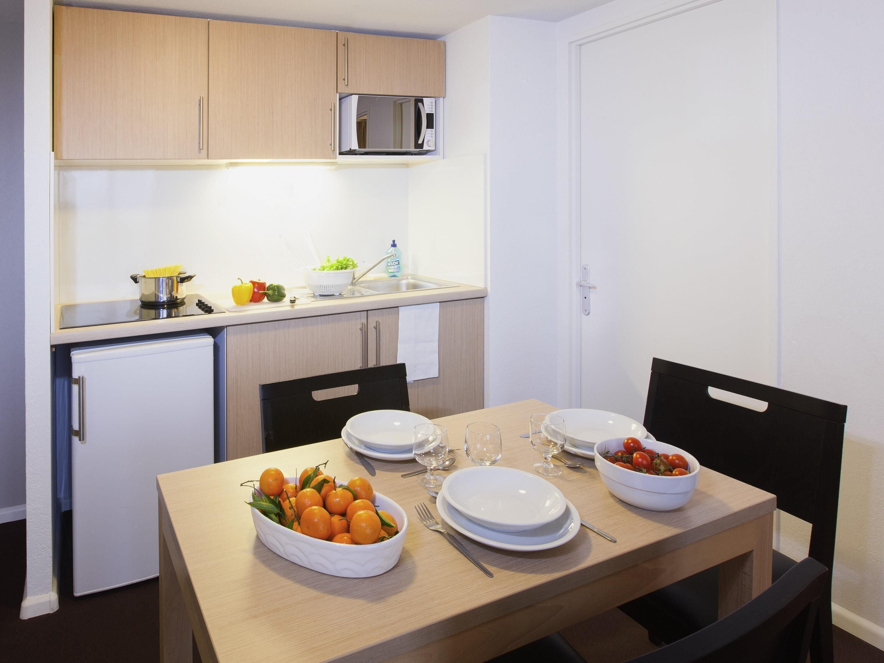 A compact kitchen with a microwave, sink, and small refrigerator. There is a dining table set with plates, glasses, and a bowl of fruit.