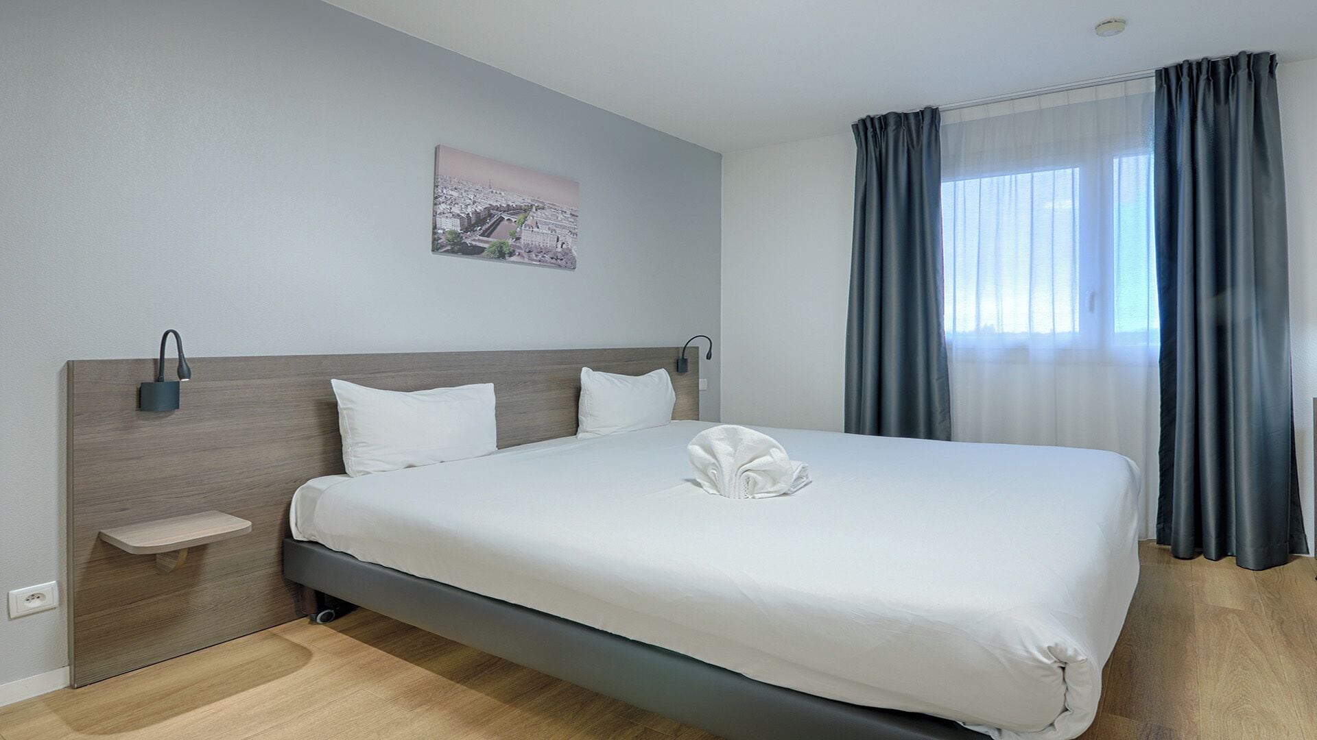 A modern hotel room with a large bed, a bedside table, a wall-mounted lamp, and a window with curtains.