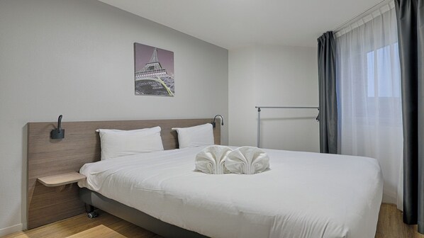 A hotel room with a large bed, a bedside table, a wall-mounted lamp, a picture of the Eiffel Tower, and a window with curtains.