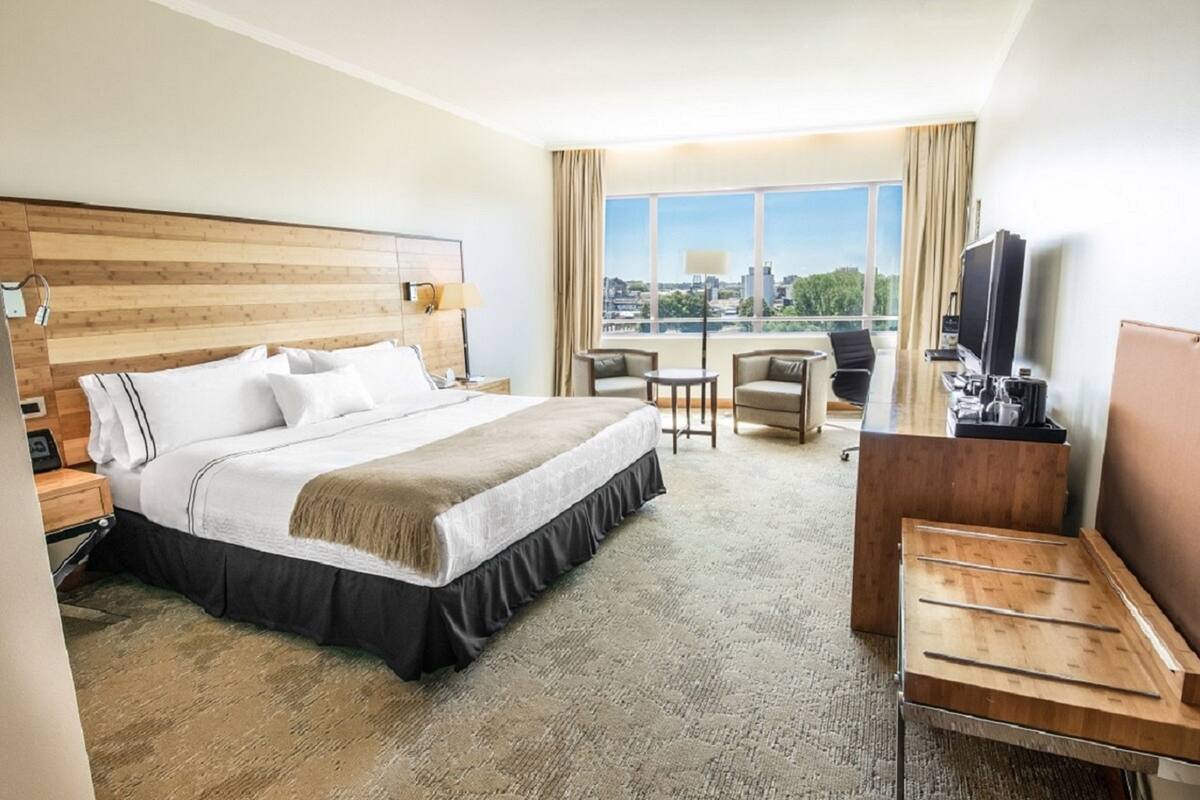 A hotel room with a bed, a desk, a chair, a TV, and a view of the city.
