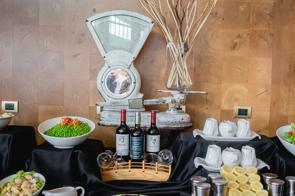 A buffet setup with a variety of food items, including a bowl of salad, a bowl of green vegetables, and a bowl of sliced fruits. There are also several bottles of wine and a tray of white teapots.