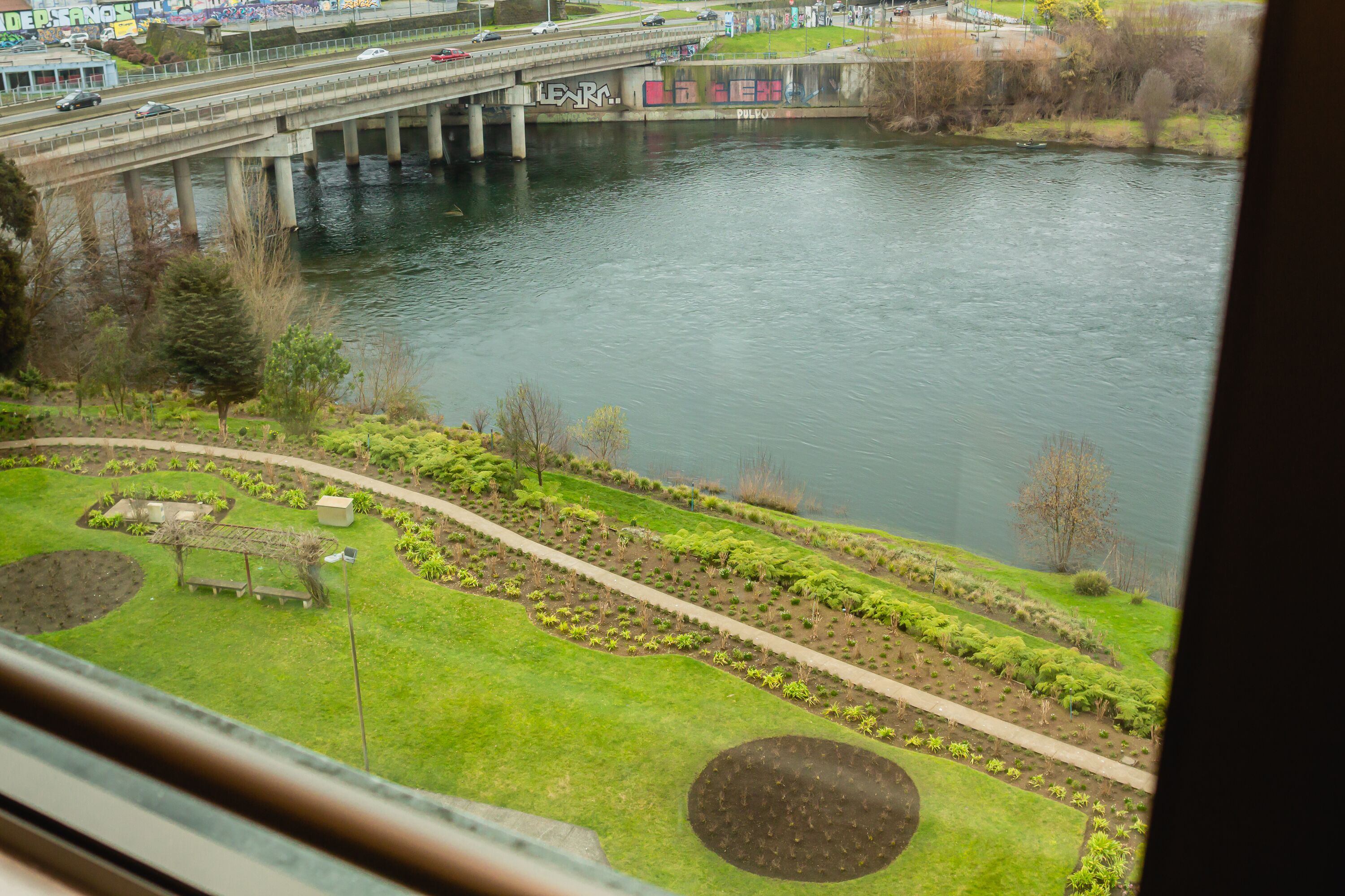 A view of a river with a bridge, a park with a walking path, and a grassy area with a circular garden bed.