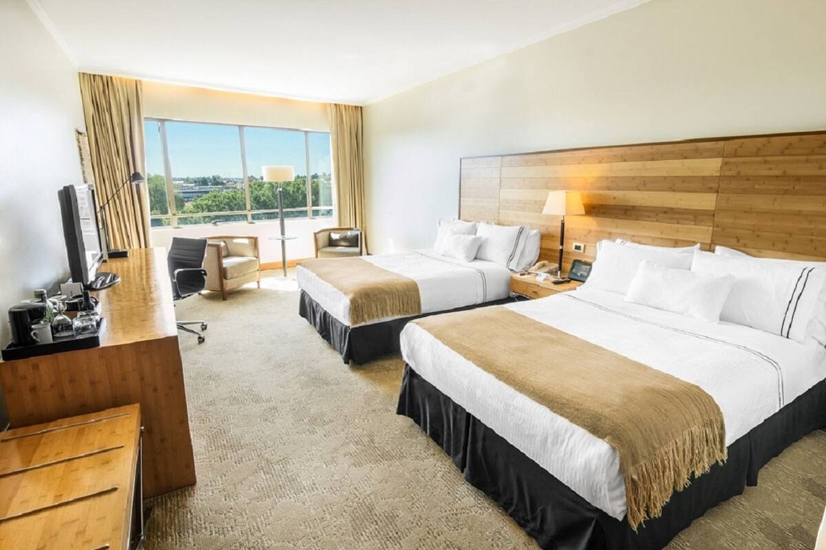 A hotel room with two beds, a desk, a chair, and a large window offering a city view.