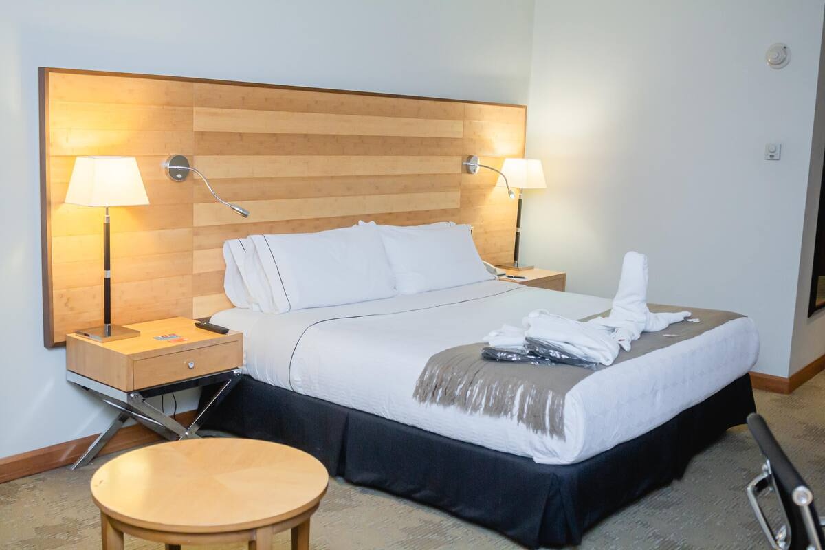 A hotel room with a wooden headboard, two bedside lamps, a bed with white linens, a round wooden table, and a black skirted bed.