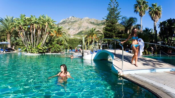 Área de piscina com água cristalina, palmeiras e uma montanha ao fundo.