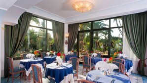Um restaurante com mesas redondas cobertas por toalhas azuis, cadeiras de madeira e grandes janelas com cortinas verdes.