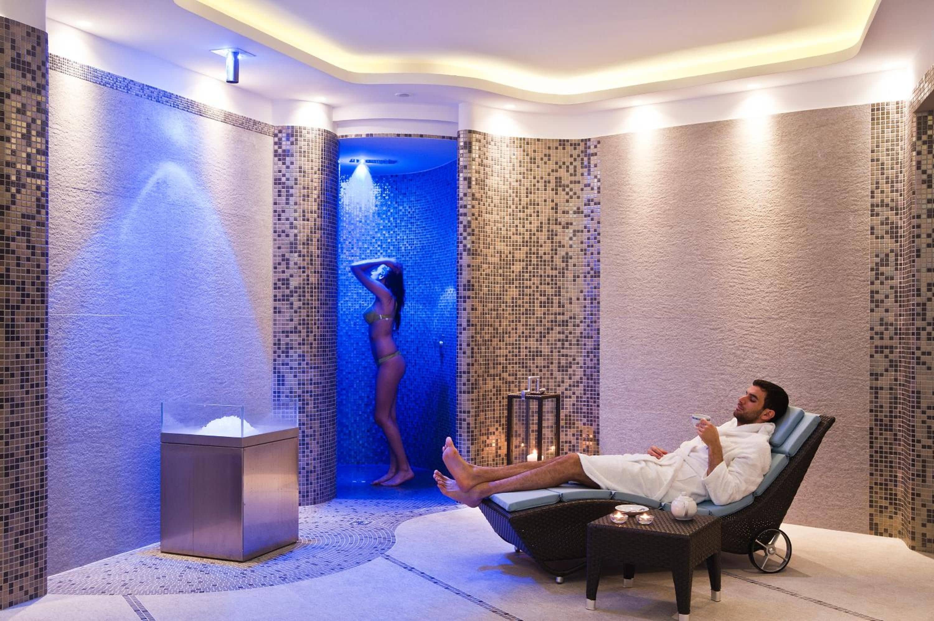 Un cuarto de spa con una persona relajándose en una silla, una mesita con un calentador de agua y una persona en la zona de la ducha.