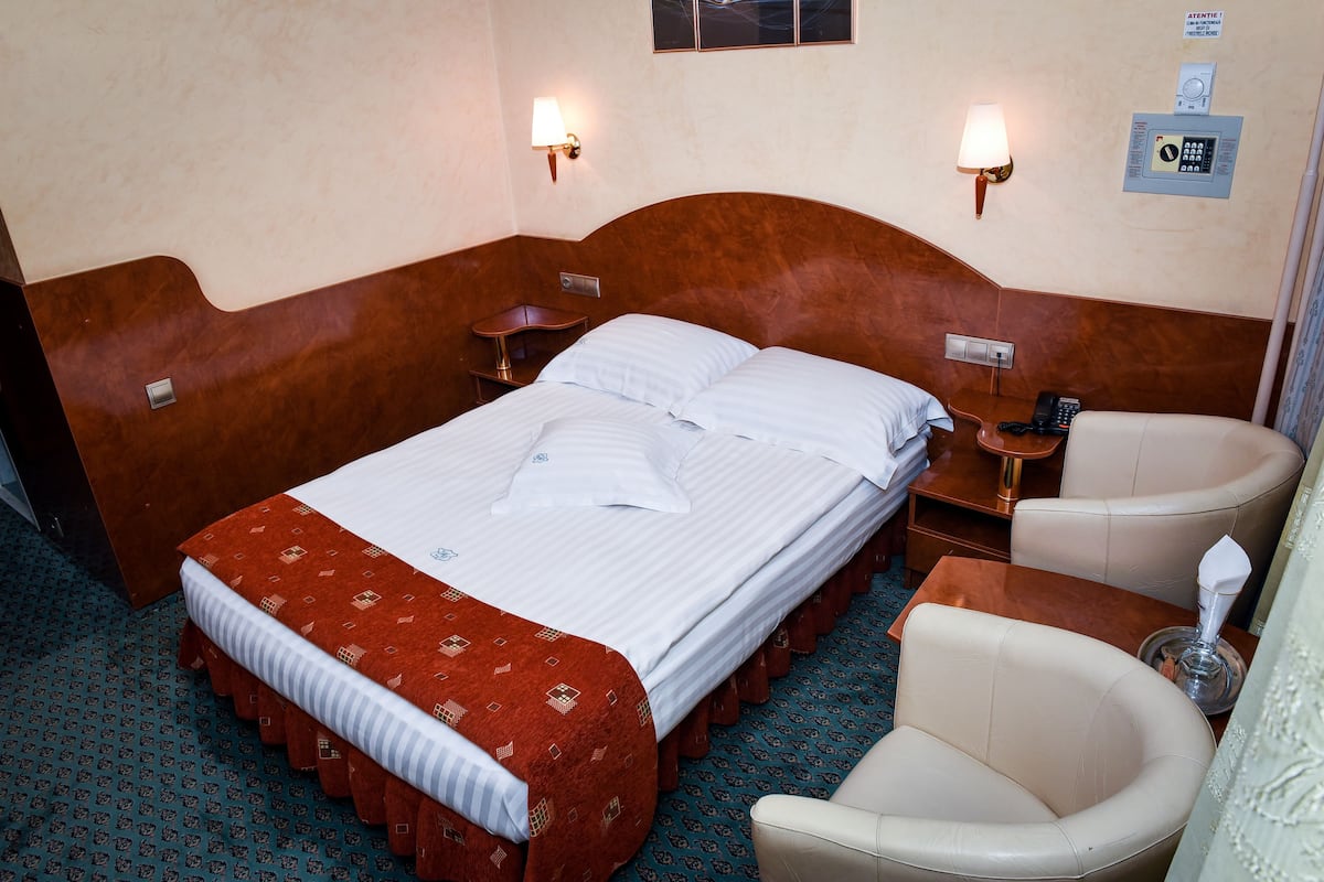 Una habitación de hotel con cabecera de madera, cama con sábanas blancas, colcha estampada roja, una mesita de noche con lámpara, una silla y un teléfono.