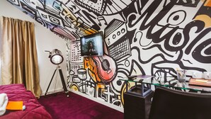 Habitació amb un mural gran a la paret, un llit amb un mantell vermell, un trípode i una televisió.