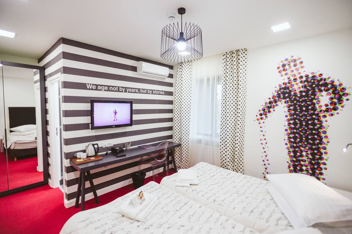 A hotel room with a striped wall, a television, a desk with a chair, a bed with a white comforter, and a colorful wall art piece.