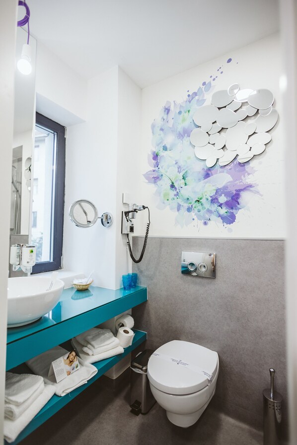 Une salle de bain moderne avec un lavabo blanc, un plan de toilette bleu et des toilettes. On y trouve un miroir, un porte-serviettes et une pomme de douche fixée au mur.