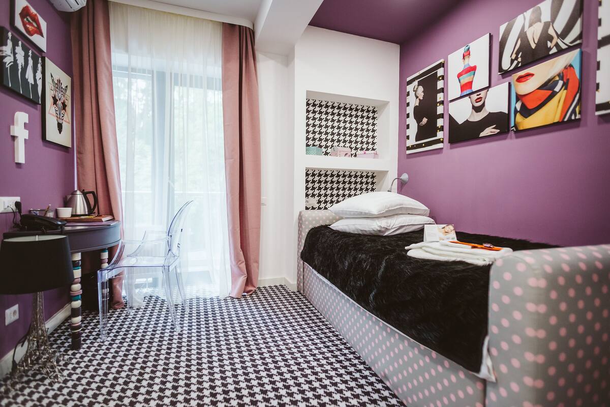 A bedroom with a black and white houndstooth floor, a bed with a black and white polka dot cover, a desk with a lamp, and a window with pink curtains.