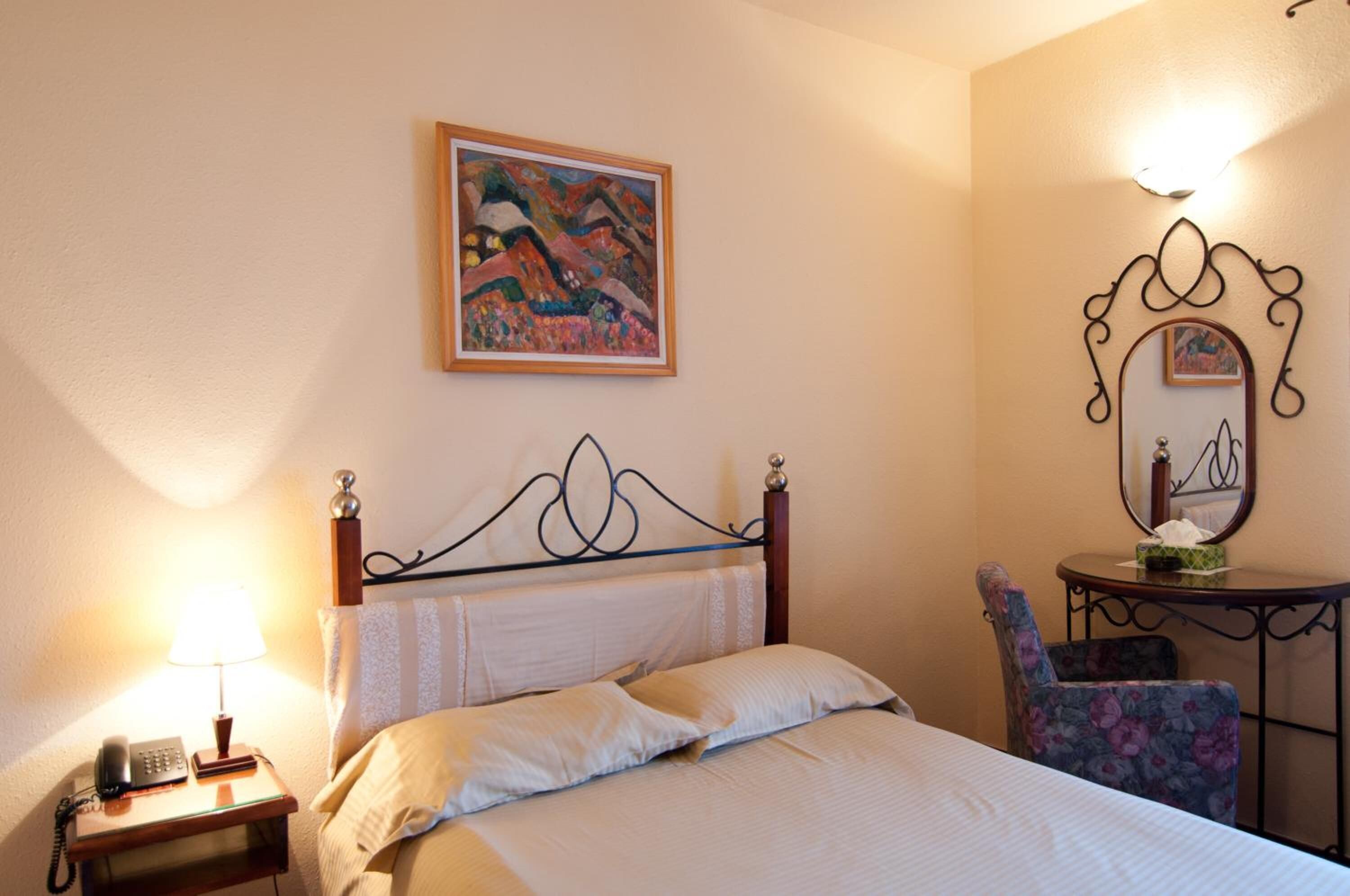 Una habitación de hotel con cama, mesita de noche con lámpara, una silla, una pequeña mesa con espejo y un cuadro en la pared.