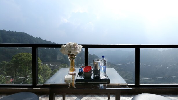 A table with a vase of flowers, a water bottle, a thermos, and a cup, set against a scenic mountain view from a balcony. - Winnies (Kasauli)