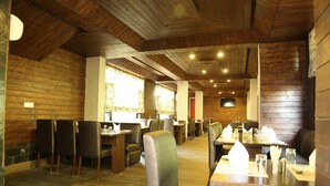 A restaurant with wooden walls and ceiling, multiple tables and chairs, and a view of the outside through large windows. - Winnies (Kasauli)