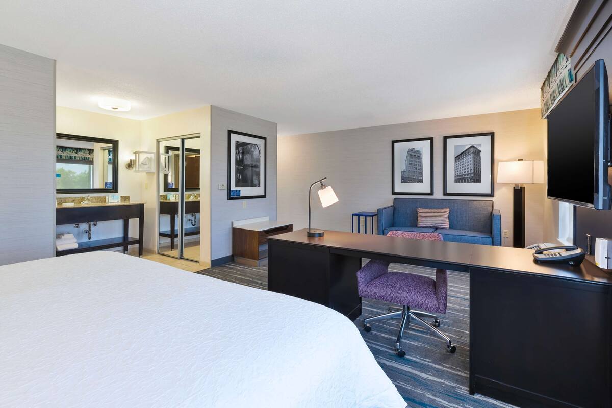 A hotel room with a large bed, a desk with a chair, a sofa, and two framed pictures on the wall.