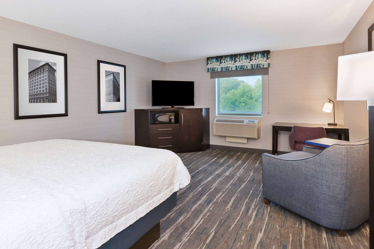 A hotel room with a bed, a television, a desk with a lamp, and framed pictures on the wall.