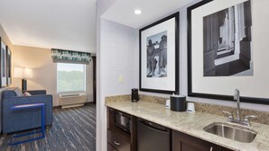 A hotel room with a kitchenette, a sofa, a window with a view, and framed artwork on the wall.