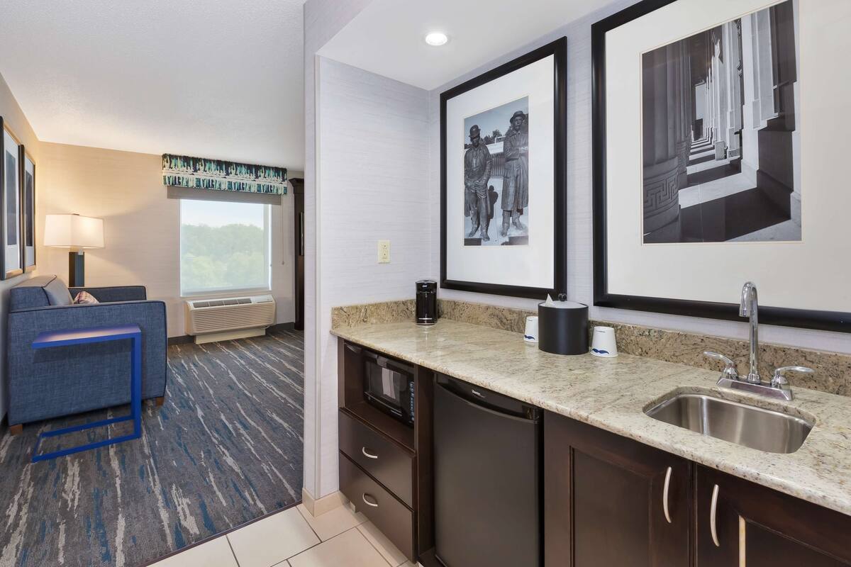 A hotel room with a kitchenette, a sofa, a window with a view, and framed artwork on the wall.