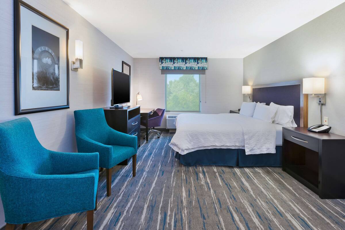 A hotel room with a bed, two chairs, a desk, a TV, and a window with a view of trees.