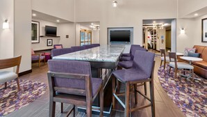 A modern hotel lobby with a long communal table, purple upholstered chairs, and a patterned rug.