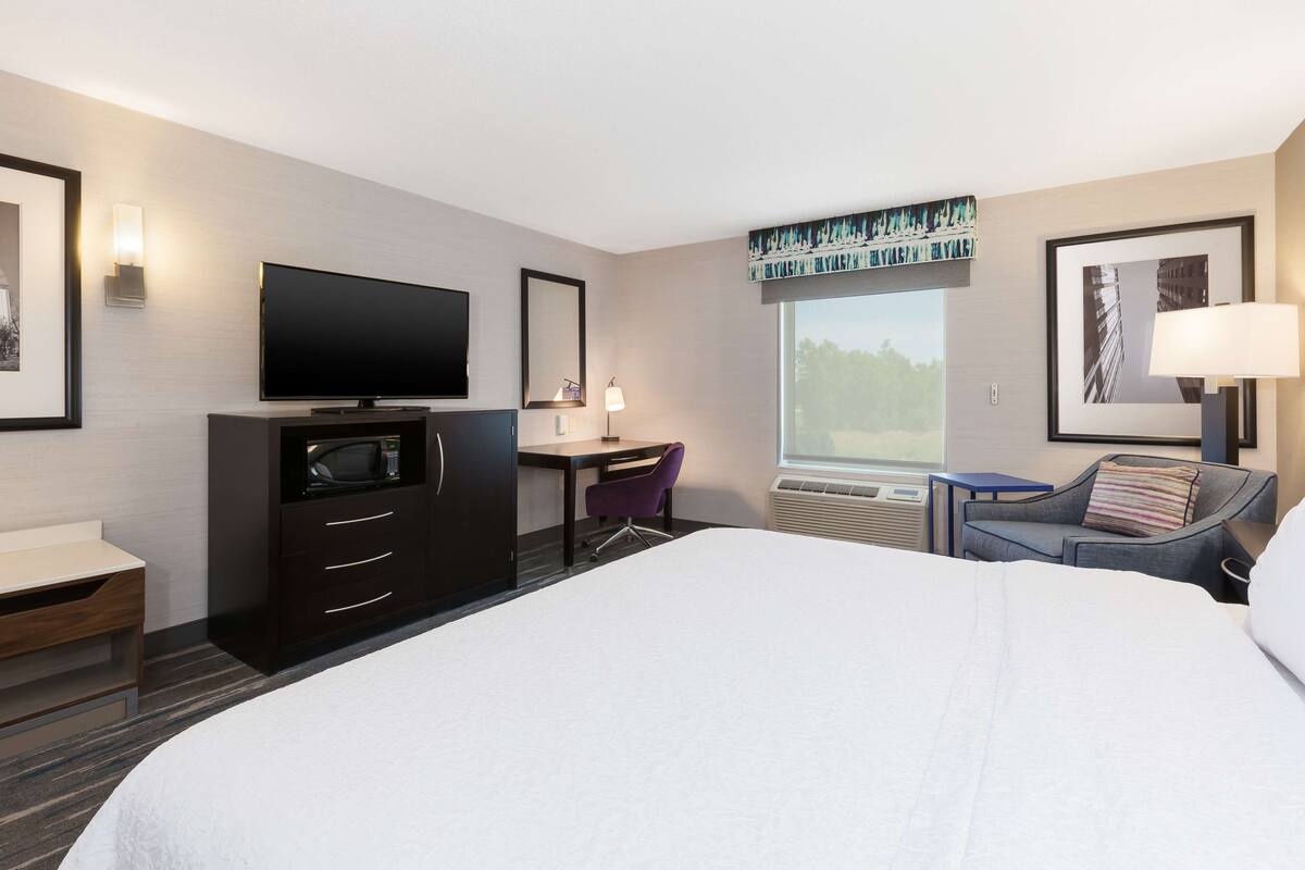 A hotel room with a large bed, a desk with a flat-screen TV, a chair, a small table, and a window with a view of trees.