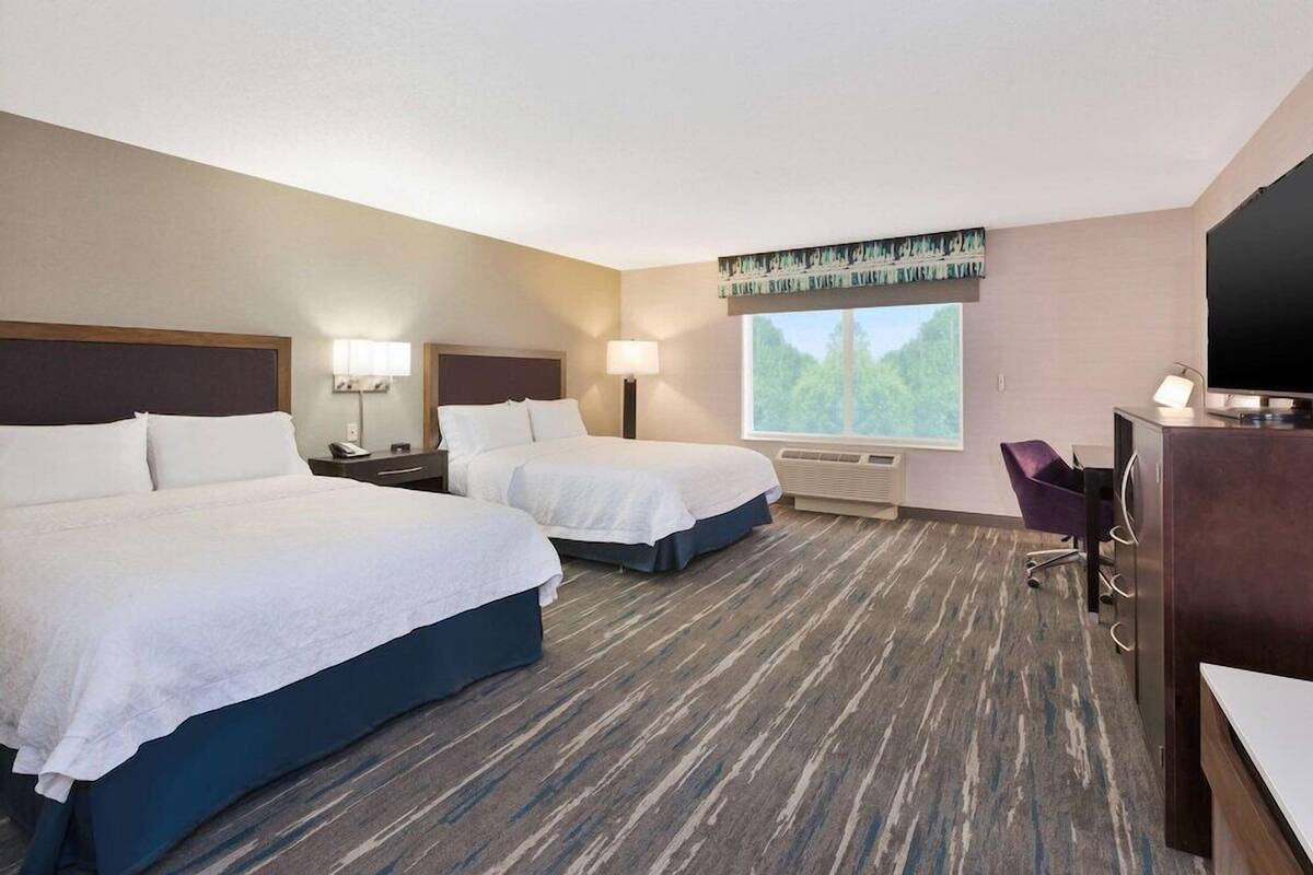 A hotel room with two beds, a desk with a computer, a chair, and a large window with a view of trees.