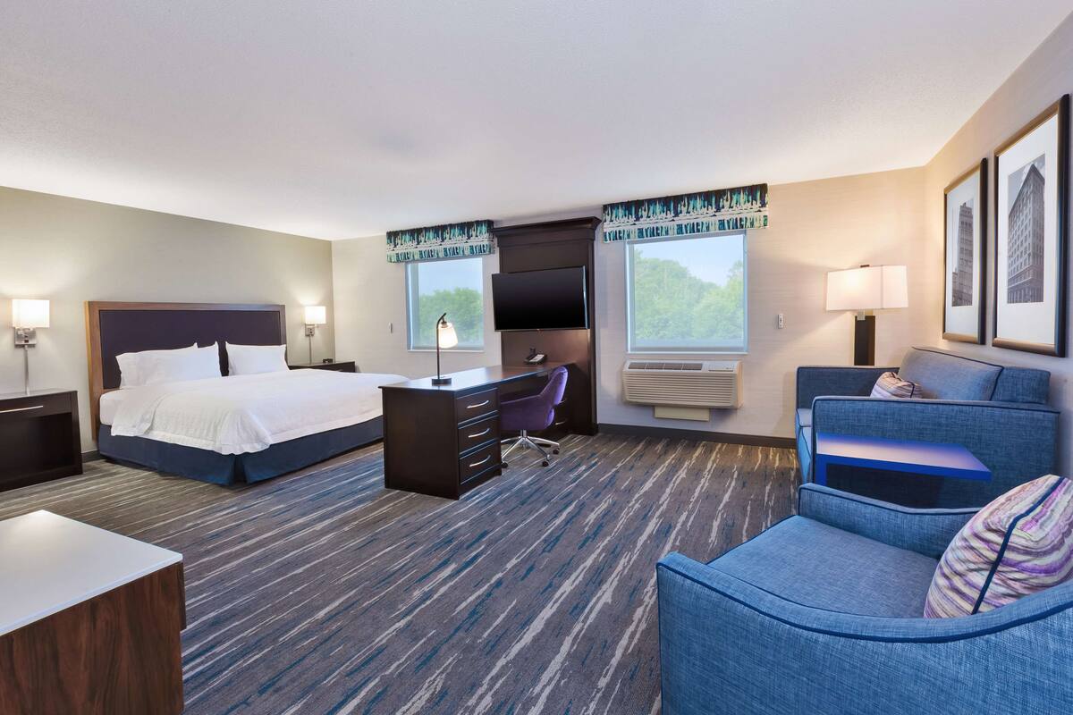 A hotel room with a bed, desk, chair, and a large window.