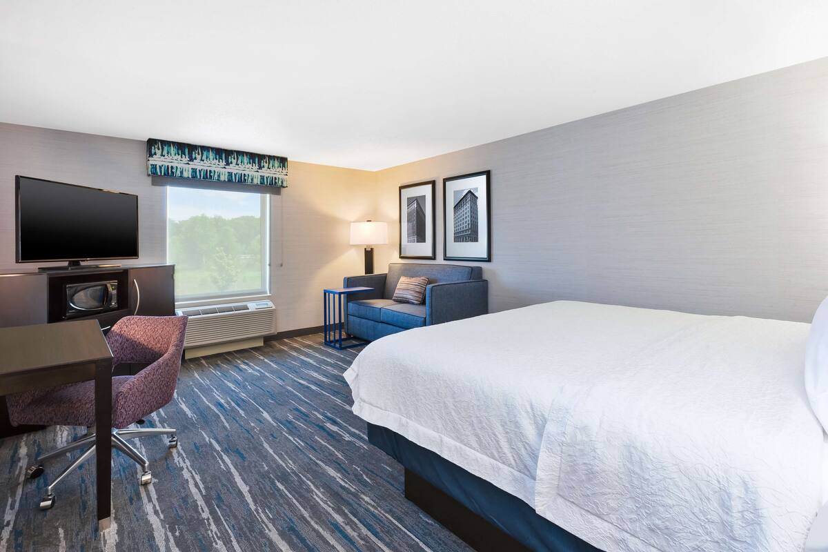 A hotel room with a large bed, a desk with a flat-screen TV, a chair, a small table, and two framed pictures on the wall.