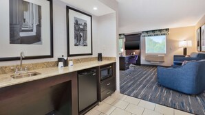A modern hotel room with a kitchen area, a seating area with a blue sofa, and a flat-screen TV.