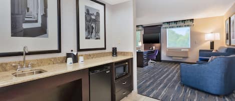 A modern hotel room with a kitchen area, a seating area with a blue sofa, and a flat-screen TV.