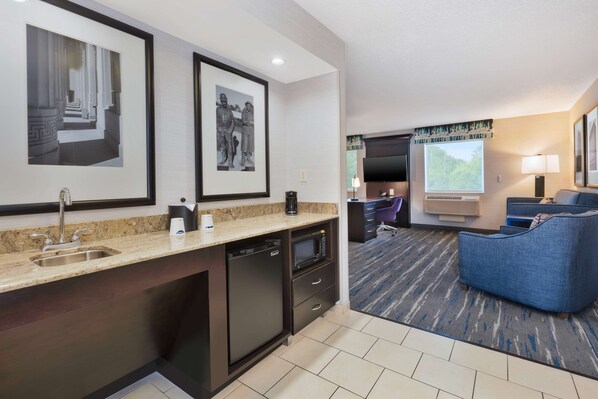 A modern hotel room with a kitchen area, a seating area with a blue sofa, and a flat-screen TV.