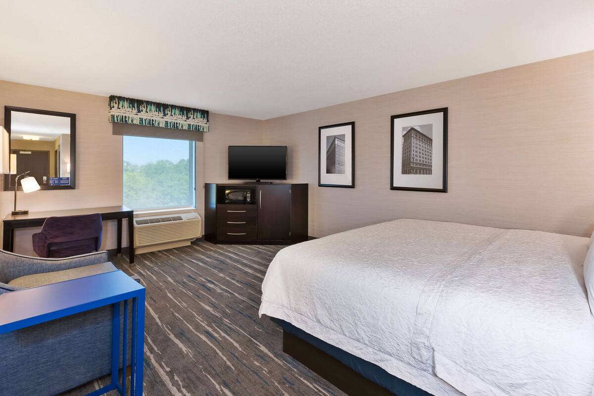 A hotel room with a large bed, a desk with a chair, a flat-screen TV, a dresser, and framed pictures on the wall.