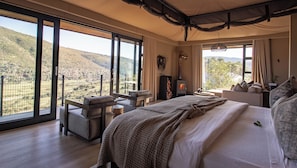 A bedroom with a large bed, a desk, and a chair, with a view of a mountainous landscape through large windows.