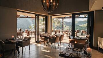 Restaurante com várias mesas e cadeiras, grandes janelas com vista para o exterior e um lustre moderno.