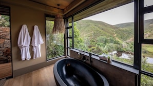 A modern bathroom with a large bathtub, a stone wall, and a view of a river and hills.