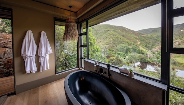 Un baño moderno con una bañera grande, una pared de piedra y vistas a un río y colinas.