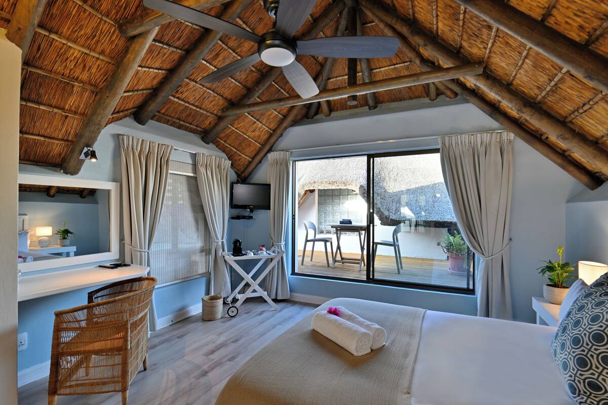 A room with a sloped ceiling, a bed with a rolled towel, a desk with a chair, a small table with chairs, and a view of an outdoor patio.